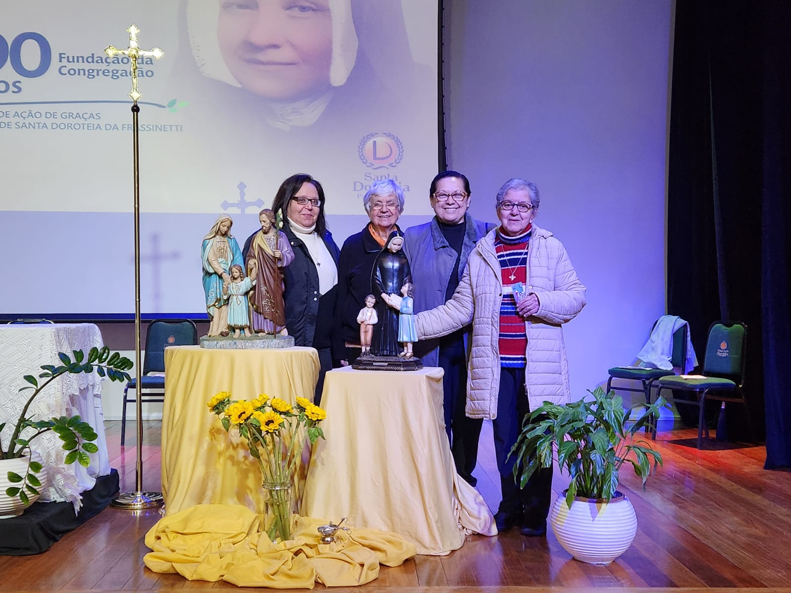 Quatro irmãs dorotéias estão uma ao lado da outra, no palco do Teatro do Colégio Santa Dorotéia, junto da imagem de Santa Paula Frassinetti.