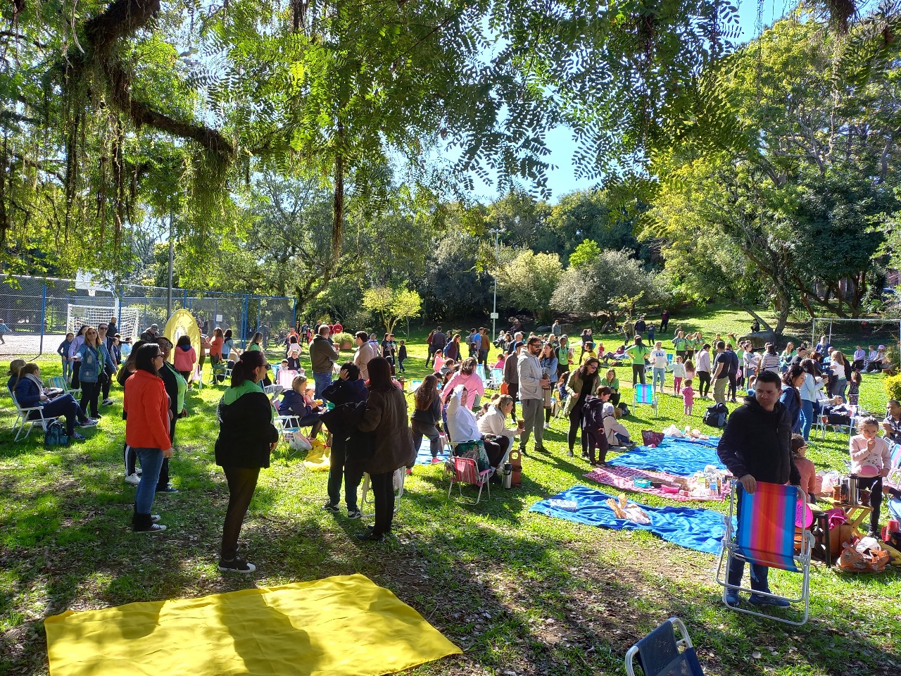 Pessoas reunidas em um praça. Elas estão sentadas em cadeiras, cangas, conversando e tomando chimarrão, em um dia de sol. Todas vestem roupas de inverno.