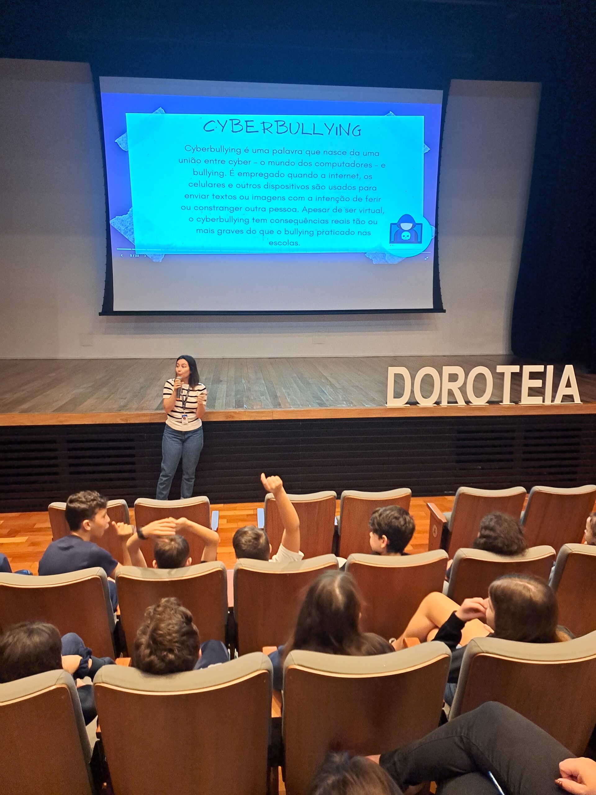 Psicóloga Ana Paula está falando para a plateia do teatro, com um microfone. Atrás dela está uma imagem de um texto com o título Cyberbullying.