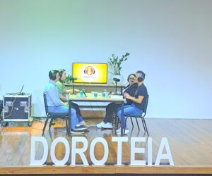 Setor de Orientação Educacional do Colégio Santa Doroteia gravam podcast. São dois homens e duas mulheres, todos brancos, em uma mesa de podcast, em cima de um placo de teatro, com um grande letreiro escrito Doroteia na freente.