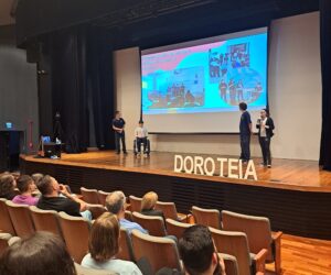 Alunos em cima do palco, com slides emitidos pelo retroprojetor, fazendo apresentação para pessoas na plateia do teatro.