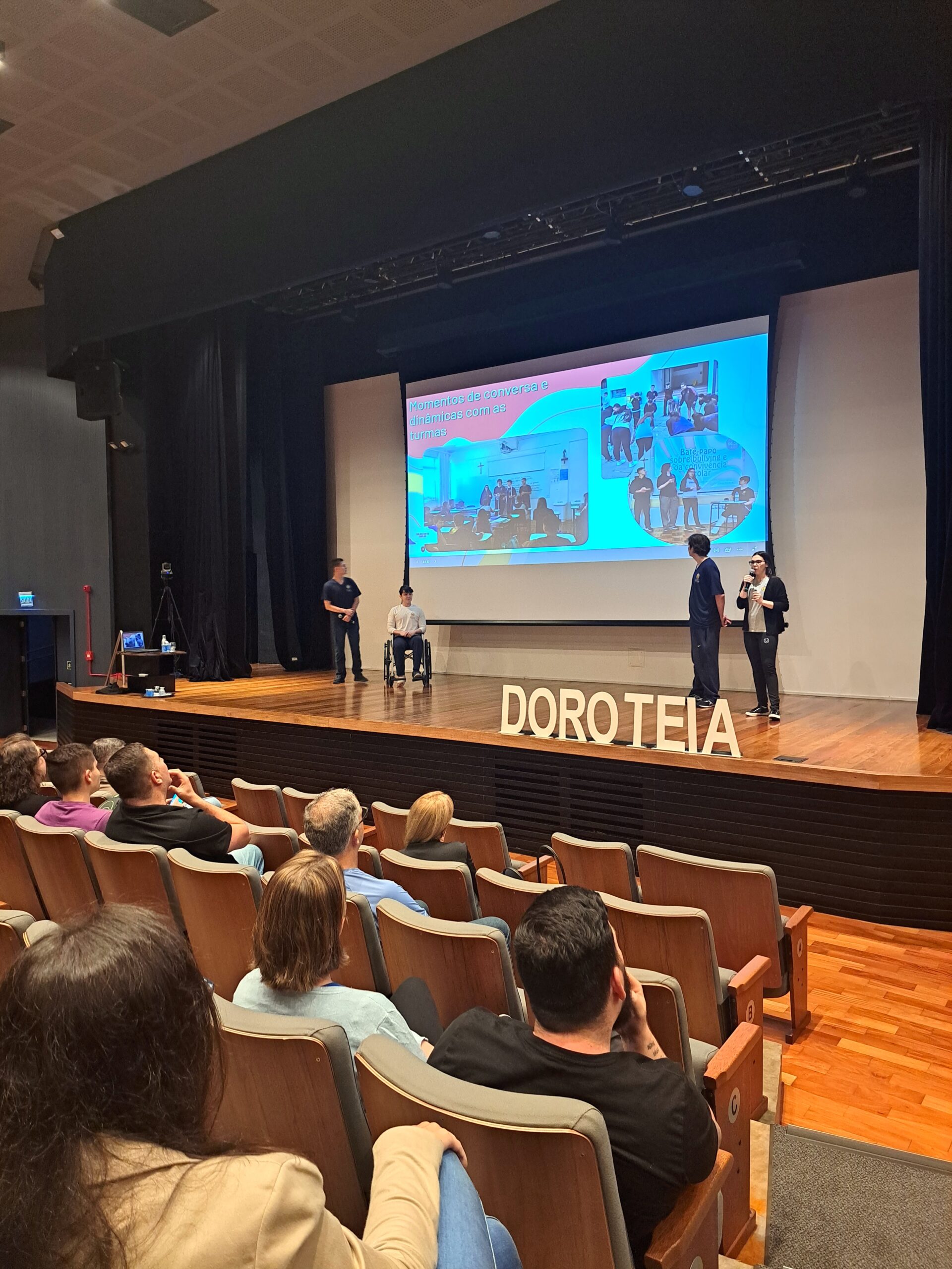 Alunos em cima do palco, com slides emitidos pelo retroprojetor, fazendo apresentação para pessoas na plateia do teatro.